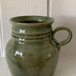 May include: A handmade, green ceramic mug with a rounded body and a handle. The mug has a slightly textured surface and three horizontal ridges near the top. The interior is a darker shade of green. The mug is suitable for hot or cold beverages.