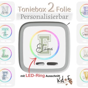 Könnte beinhalten: Ein weißer, quadratischer Aufkleber für die Toniebox mit einem zentralen Regenbogenkreis, einem großen Blockbuchstaben E und dem Namen Elias in Schreibschrift. Das Design enthält einen Ausschnitt für den LED-Ring und acht kleine Namensaufkleber.