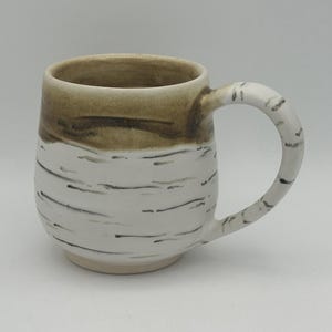 May include: A handmade ceramic mug with a birch tree design. The top of the mug is brown, transitioning to white with black horizontal lines. The handle is white with black markings. A unique, rustic coffee mug.