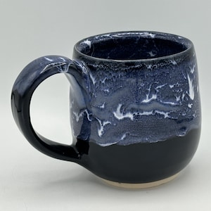 May include: A handmade ceramic mug with a rounded shape and a large handle. The mug features a two-tone design, with a glossy black base and a marbled blue and white upper section. The interior is dark blue.