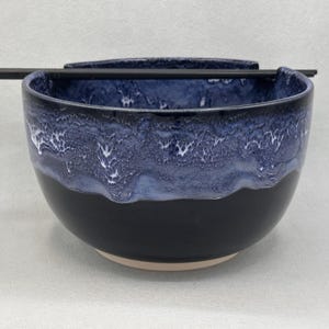 May include: A ceramic ramen bowl with a glossy black base and a textured, dripped blue and white glaze around the upper rim. The bowl features a notched edge designed to hold a pair of black chopsticks, which are resting horizontally across the top.
