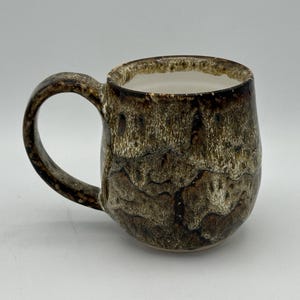 May include: A handmade ceramic mug with a unique, textured glaze in shades of brown and tan. The mug has a rounded body, a sturdy handle, and a white interior. The exterior features a marbled effect, adding to its rustic charm.