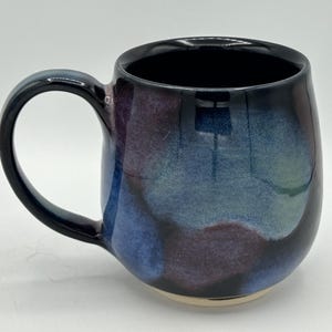 May include: A ceramic mug with a rounded body and a large handle. The mug features a glossy finish with a blend of blue, purple, and green hues. The interior and handle are black, and the base is unglazed.
