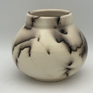 May include: A handcrafted, off-white ceramic vase with a rounded body and a slightly flared rim. The vase features unique black, lightning-like patterns across its surface, adding a striking contrast to the neutral background. This decorative piece is ideal for home decor.