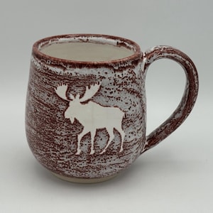 May include: A ceramic mug with a white moose silhouette on a textured, reddish-brown background. The mug has a rounded shape, a large handle, and a white interior. The mug is suitable for hot beverages.