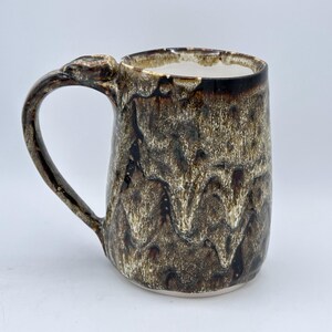 May include: A tall ceramic mug with a textured, dripped glaze in shades of brown and cream. The mug has a rounded handle and a white interior. The glaze pattern gives the mug a unique, handcrafted appearance.