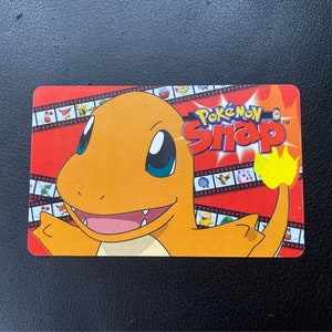 May include: A rectangular Pokemon Snap card featuring Charmander, an orange lizard-like creature with a flame on its tail. The card has a red background with the words "Pokemon Snap" and film strip-like borders.