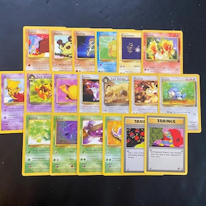 May include: A collection of Pokémon trading cards, featuring various characters with yellow borders. The cards display character names like Diglett, Mankey, and Abra, along with game-related text and illustrations. The cards are arranged on a dark surface.