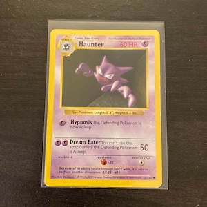 May include: A Haunter Pokemon card from the 1999 Wizards of the Coast set. The card is yellow with a purple and black illustration of Haunter. The card text reads "Hypnosis The Defending Pokemon is now Asleep." and "Dream Eater You can't use this attack unless the Defending Pokemon is Asleep. 50". The card also has a weakness, resistance, and retreat cost.