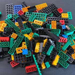 May include: A large collection of assorted plastic building blocks in various shapes and sizes. The pile includes rectangular, square, and flat pieces in vibrant colors such as black, green, yellow, red, and blue, featuring circular studs.