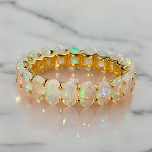 May include: A gold eternity band featuring a continuous row of oval-shaped opals. Each opal displays iridescent flashes of green, blue, and orange colour. The stones are set in a polished gold metal frame that encircles the entire circumference.