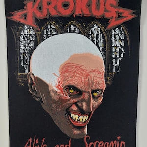 May include: Black patch featuring the band name "Krokus" in red, above a grotesque face with yellow eyes and a sinister grin. The text "Alive and Screamin" is below the face. The background includes a church window design.