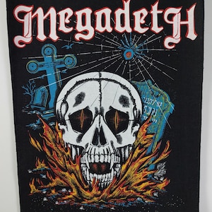 May include: Black patch with a skull design, orange flames, and the word "Megadeth" in white with a red outline. The design includes a spiderweb, a cross, and a tombstone.
