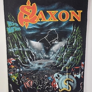 Peut inclure: Écusson rectangulaire noir avec le nom du groupe "Saxon" en orange et jaune. L'écusson représente une scène de concert avec une foule, des drapeaux, des montagnes et un ciel orageux. Le texte "R&B The Nations" est également visible.
