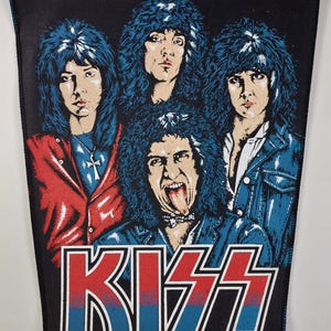 Könnte beinhalten: Schwarzer rechteckiger Aufnäher mit einem Porträt der Rockband KISS. Die Bandmitglieder haben blaue Haare und tragen Jacken. Der Name der Band, "KISS", ist in roten und blauen Blockbuchstaben dargestellt.