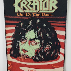 Puede incluir: Un parche negro con el nombre de la banda "Kreator" en letras verdes y naranjas. Debajo, una ilustración de una cara de zombi en un charco de líquido rojo, con el texto "Out Of The Dark..." debajo del nombre de la banda. Borde cosido.