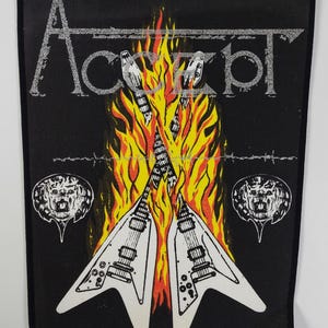 May include: Black patch with the band name "Accept" in silver lettering. The design features two white guitars with flames, two lion head illustrations, and barbed wire details.