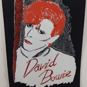 May include: A black rectangular patch featuring a stylized portrait of David Bowie with red hair and red lips. The name "David Bowie" is written in red script below the portrait. The background is black with a white and red design.