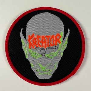 May include: Embroidered patch featuring a gray alien-like face with green accents and the word "KREATOR" in orange across the forehead. The patch has a black background with a red border.