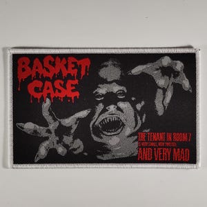 Peut inclure: Écusson rectangulaire noir avec une bordure blanche. L'écusson présente les mots "BASKET CASE" en lettres rouges dégoulinantes au-dessus d'un visage grotesque et hurlant avec les mains tendues. En dessous, le texte indique "THE TENANT IN ROOM 7 IS VERY SMALL, VERY TWISTED AND VERY MAD."