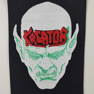 Puede incluir: Parche negro con un diseño de rostro verde y blanco estilizado con orejas puntiagudas. La palabra "KREATOR" está impresa en rojo en la frente. El parche tiene un borde negro.
