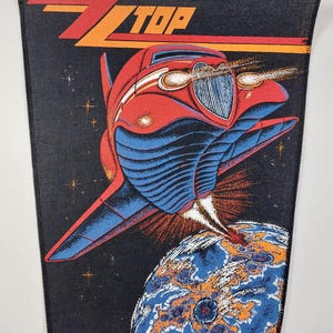 Peut inclure: Écusson noir avec le nom du groupe ZZ Top en orange et rouge. L'écusson représente une voiture hot rod rouge et bleue décollant d'une planète. Le fond est noir avec des étoiles.