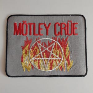 May include: Embroidered patch featuring the band name "Mötley Crüe" in red, above a pentagram encircled by flames. The patch has a gray background and a black border, suitable for jackets or bags.