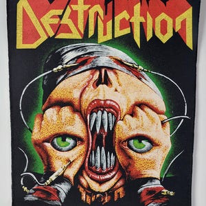 Może przedstawiać: Czarna naszywka z napisem "Destruction" w żółtych i czerwonych literach blokowych. Grafika przedstawia czaszkę z odsłoniętymi zębami i dłońmi zakrywającymi oczy, z zielonymi oczami i zielonym tłem.