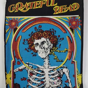Puede incluir: Parche negro con las palabras "Grateful Dead" en amarillo y rojo. El diseño presenta un esqueleto con una corona de rosas rojas sobre un fondo azul, enmarcado por un borde decorativo rojo y amarillo.