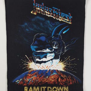 Puede incluir: Parche rectangular negro con el nombre de la banda "Judas Priest" en una fuente multicolor. El parche representa un puño robótico azul y negro golpeando hacia abajo, con las palabras "RAM IT DOWN" en dorado.