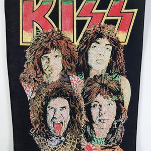 Könnte beinhalten: Schwarzer rechteckiger Aufnäher mit der Band KISS. Der Name der Band steht in großen roten Buchstaben mit gelber Umrandung oben. Darunter befinden sich vier Porträts der Bandmitglieder mit langen Haaren und Make-up.