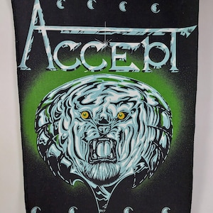 May include: Black patch featuring the word "ACCEPT" in white and teal lettering. Below the text is a detailed illustration of a snarling tiger with teal and white stripes and yellow eyes, set against a green and black background. Crescent moon shapes are at the top and bottom.