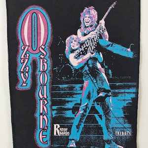 Op de afbeelding: Zwarte stoffen patch met een levendige illustratie van Ozzy Osbourne en Randy Rhoads op het podium. Het ontwerp bevat de tekst "Ozzy Osbourne" in roze en blauw, met "Randy Rhoads" en "Tribute" ook zichtbaar. De afbeelding toont een dynamische rock and roll-performance.