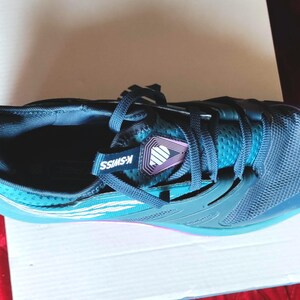 K&ouml;nnte beinhalten: Ein Paar blaue und t&uuml;rkisfarbene Sportschuhe mit einem wei&szlig;en K-Swiss-Logo an der Seite. Die Schuhe haben ein schwarzes Mesh-Obermaterial und eine wei&szlig;e Sohle.