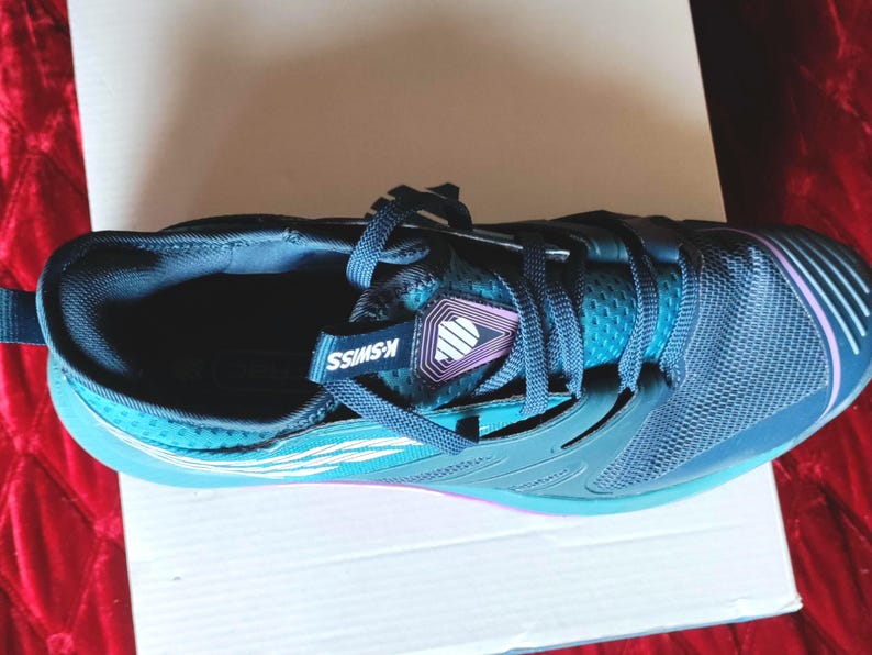 K&ouml;nnte beinhalten: Ein Paar blaue und t&uuml;rkisfarbene Sportschuhe mit einem wei&szlig;en K-Swiss-Logo an der Seite. Die Schuhe haben ein schwarzes Mesh-Obermaterial und eine wei&szlig;e Sohle.