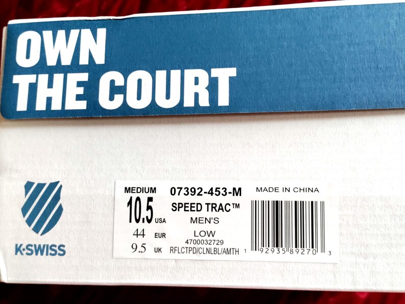 K&ouml;nnte beinhalten: Ein wei&szlig;er Schuhkarton mit den Worten "OWN THE COURT" in Blau auf der Oberseite. Der Karton ist mit dem Markennamen "K-SWISS" und der Schuhgr&ouml;&szlig;e "10.5" in US-Gr&ouml;&szlig;en gekennzeichnet. Das Schuhmodell ist "SPEED TRAC" und der Schuh wird als "LOW" und "MEN'S" beschrieben. Der Karton enth&auml;lt auch einen Barcode und den Text "MADE IN CHINA".
