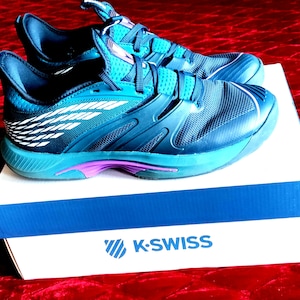 K&ouml;nnte beinhalten: Ein Paar blaue und t&uuml;rkisfarbene K-Swiss Tennisschuhe mit wei&szlig;en Akzenten auf einem wei&szlig;en Karton mit dem K-Swiss Logo.