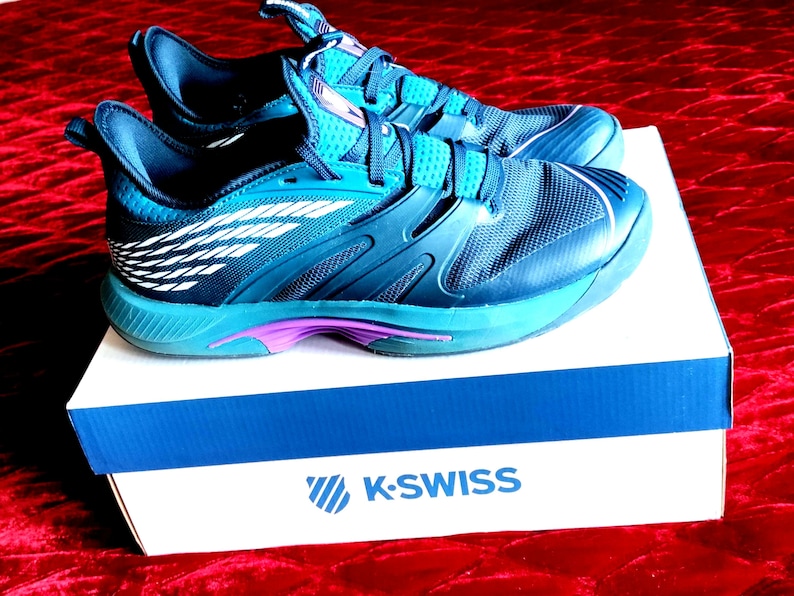 K&ouml;nnte beinhalten: Ein Paar blaue und t&uuml;rkisfarbene K-Swiss Tennisschuhe mit wei&szlig;en Akzenten auf einem wei&szlig;en Karton mit dem K-Swiss Logo.