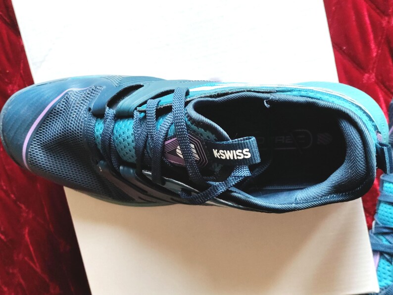 K&ouml;nnte beinhalten: Ein Paar blaue und t&uuml;rkisfarbene Sportschuhe mit einem wei&szlig;en K-Swiss-Logo an der Seite. Die Schuhe haben ein Mesh-Obermaterial und eine schwarze Sohle.