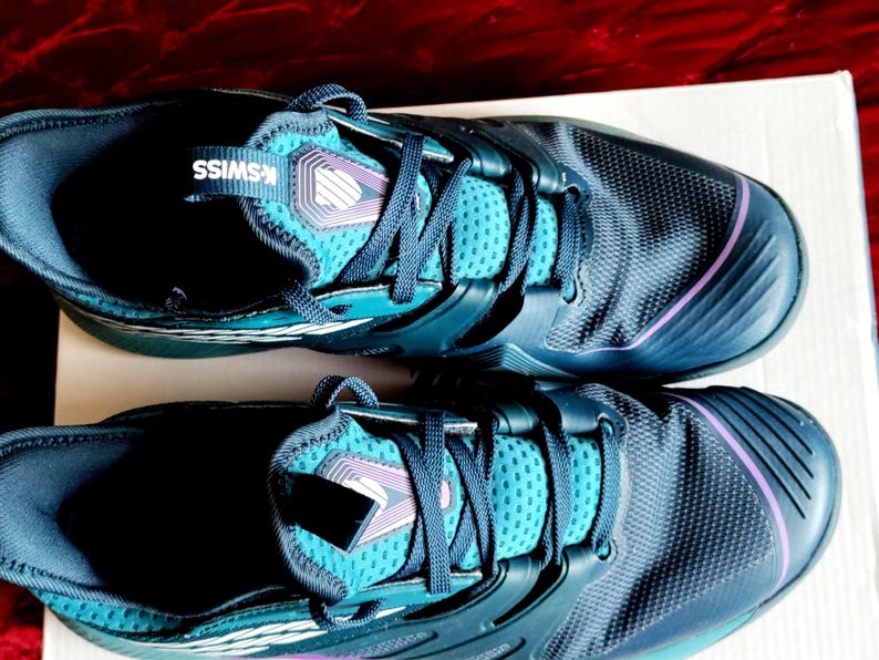 K&ouml;nnte beinhalten: Ein Paar dunkelblaue Sportschuhe mit t&uuml;rkisfarbenen Akzenten und einem wei&szlig;en K-Swiss-Logo. Die Schuhe haben ein strukturiertes Obermaterial und einen violetten Besatz.
