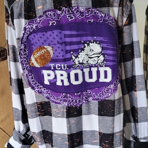 May include: A black and white plaid flannel shirt with a purple patch on the back. The patch features a football, a mascot, and the text "TCU PROUD".