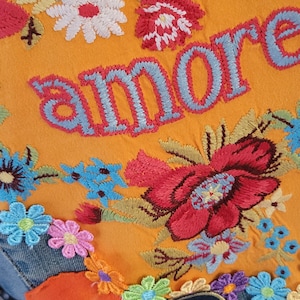May include: A denim jacket with embroidered flowers and the word "amore" in blue and red thread. The jacket has a patch with the word "question" on it and a smiley face patch with a rainbow.