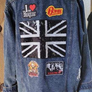 May include: A blue denim jacket with various band patches, including "I Love The Beatles", "Bowie", "Queen", and "The Rolling Stones". The jacket also features a sequined Union Jack patch.