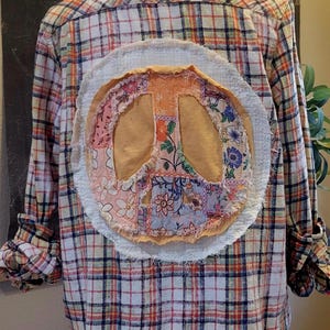 May include: A long-sleeve button-down shirt featuring a white, orange, and blue plaid pattern. The back displays a large, circular, frayed-edge applique patch with a peace sign design made from various floral and solid-colored fabric scraps.