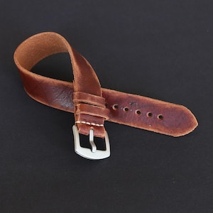 May include: A brown leather watch strap with a silver buckle. The strap has a distressed look and is folded in half.