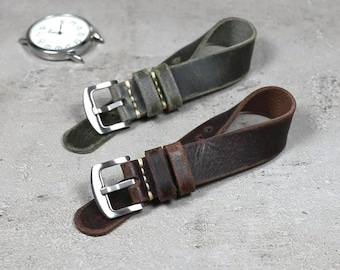 May include: Two leather watch straps in shades of brown and green with silver buckles. The straps are shown on a gray surface with a silver watch in the background. The watch straps are a vintage style.
