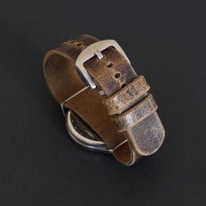 May include: A brown leather watch strap with a silver buckle. The strap has a distressed look, with a worn and faded appearance.