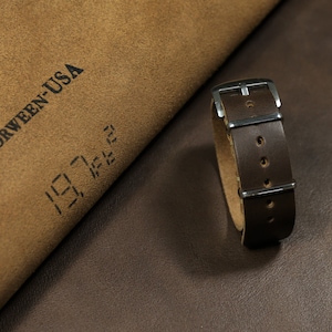 Könnte beinhalten: Ein braunes Lederarmband mit silberner Hardware. Das Armband ist aus Horween-Leder gefertigt, einem hochwertigen Leder, das für seine Langlebigkeit und Strapazierfähigkeit bekannt ist. Das Armband wird auf einer braunen Lederoberfläche gezeigt.