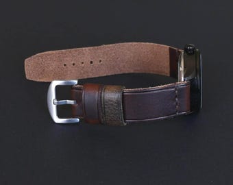 May include: A close-up of a brown leather watch with a silver buckle. The watch band is a dark brown color, and the watch face is black. The watch is set against a dark background.