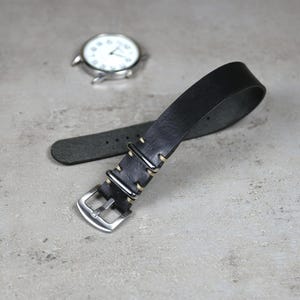May include: A black leather watch strap with a silver buckle and metal loops. The strap is lying on a gray surface, with a silver watch in the background.